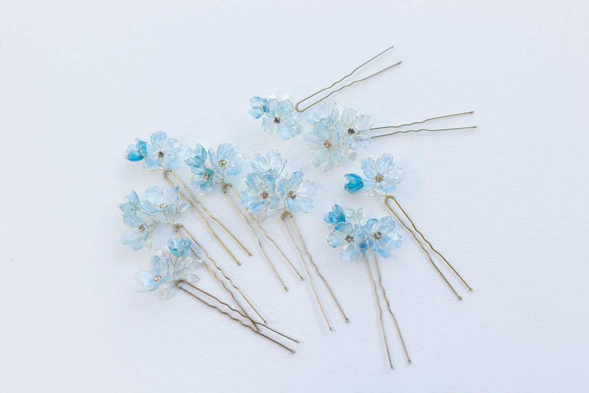 Translucent blue flower bridal hair pins arranged diagonally on a white background, showing their two-prong shape.