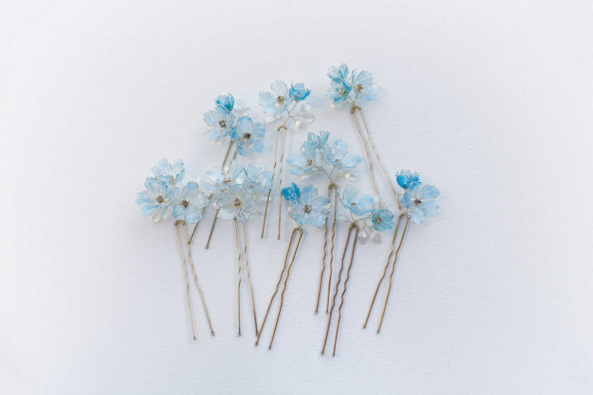 Nine blue floral bridal hair pins on a white background, each with a different arrangement of translucent flowers.