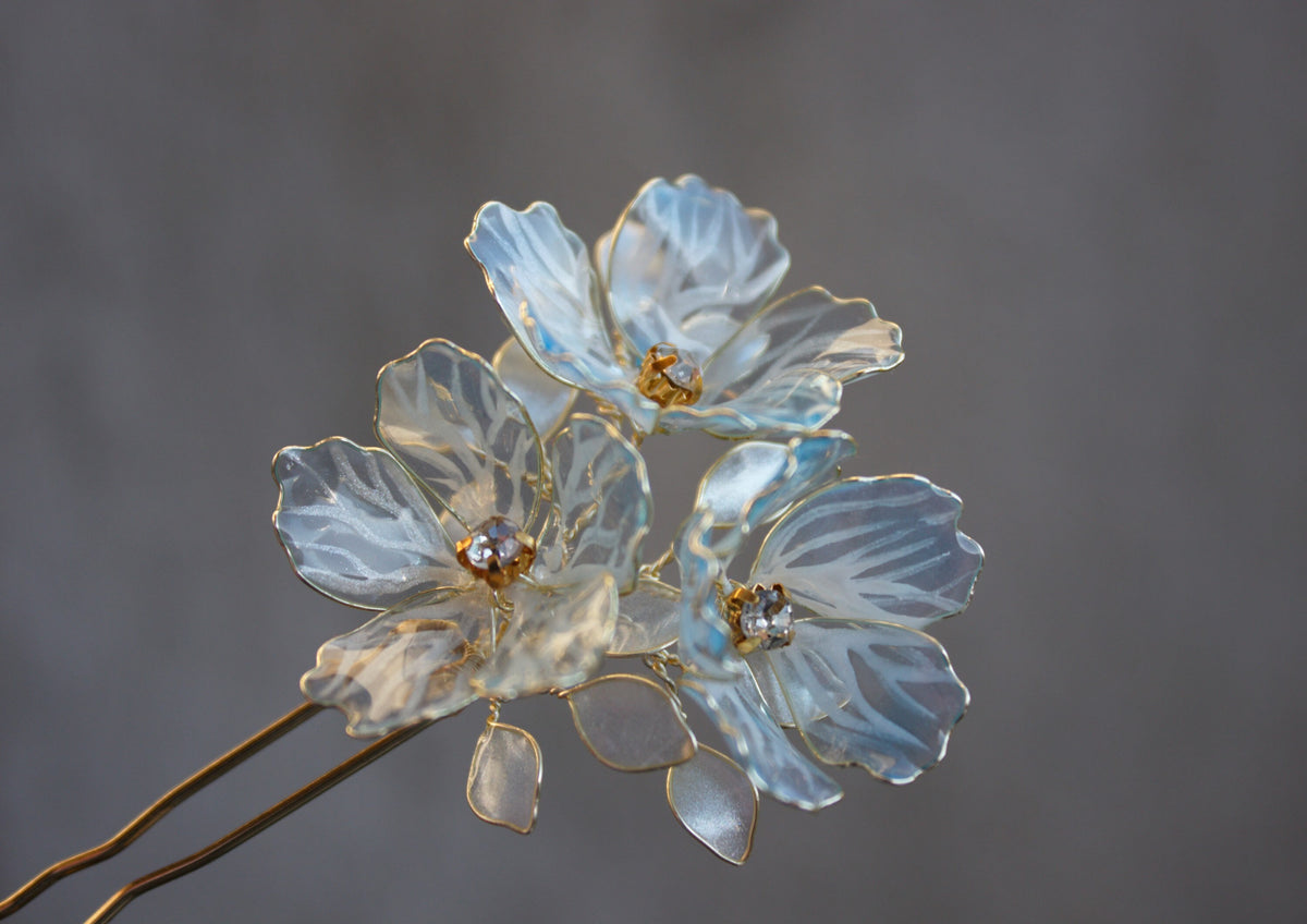 Blue floral bridal hair fork with three translucent blossoms and two metal prongs against a grey background.